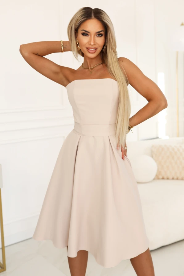 Modelka prezentuje elegancką jasnobeżową sukienkę midi bez ramiączek z rozkloszowanym dołem i podkreśloną talią, idealną na wesele, komunię i przyjęcie.