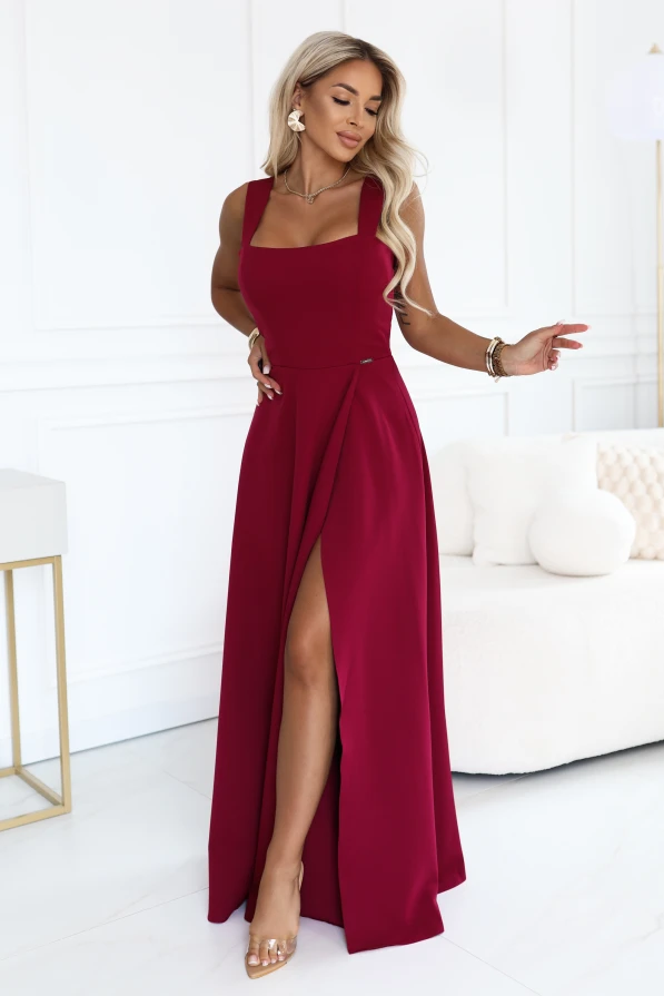 Modelka w eleganckiej bordowej sukience maxi Numoco z kwadratowym dekoltem, szerokimi ramiączkami, dopasowaną talią i wysokim rozcięciem na nodze, prezentowana w jasnym, nowoczesnym wnętrzu.